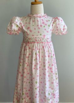 "The \"Sophie Dess\" label is usually indicative of classic clothing and fine materials and construction. This dress is a fine example. Made of a luxurious silky cotton, the print is of pink and rose flowers and buds with leaves in pale and kiwi green. The front bodice is plain without shaping and ends in a tiny self piping.  Below, at waist, is a panel of smocking done in white, pink and rose floss. The skirt below is full and gathered with two wide tucks just above hem. It will probably be mid Pink Smocked Short Sleeve Dress For Garden Party, Pink Fitted Dress With Smocked Cuffs, Fitted Classic Smocked Dress, Classic Dress With Smocked Cuffs, Pink Fitted Smocked Dress With Short Sleeves, Classic Fitted Dress With Smocked Cuffs, Fitted Cotton Smocked Dress, Pink Smocked Dress With Smocked Cuffs For Daywear, Pink Cotton Smocked Dress With Short Sleeves