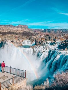 two people are standing at the edge of a waterfall with a rainbow in the background