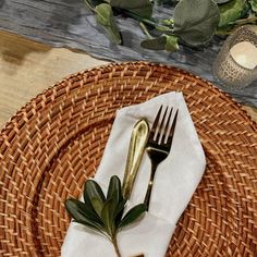 a place setting with a napkin, fork and knife on a wicker plate next to greenery