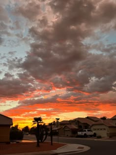 the sun is setting over some houses and palm trees in the foreground, with clouds above them