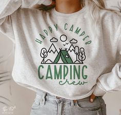 a woman wearing a happy camper sweatshirt with her hands on her head and trees in the background