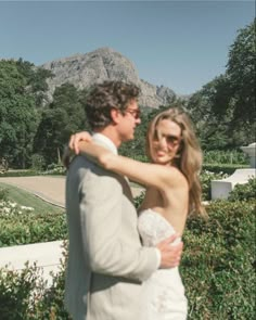 a man and woman are standing together in front of some bushes with mountains in the background