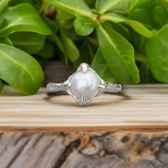 a white pearl ring sitting on top of a wooden table next to green leaves and greenery