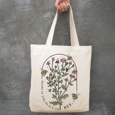 a hand holding a tote bag with flowers and bees printed on the front, against a gray background