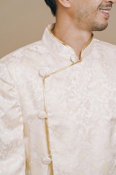 A Vietnamese ao gam jacket fit for a modern groom. This men’s Vietnamese wedding outfit features an elegant and refined design made with ivory and light gold dragon brocade fabric. Pair this with one of our beautiful ao dais for the perfect modern Vietnamese-American wedding tea ceremony. Bride's Dress Recommendation: Pairs well with our white and gold Kathy Vietnamese Wedding Dress. This jacket is named after the Tran Dynasty that ruled between 1225-1400, during which time they resisted invader Elegant Nehru Jacket For Traditional Ceremonies, Gold Brocade Outerwear For Wedding, Gold Long Sleeve Bandhgala For Traditional Ceremonies, Elegant Traditional Wear With Stand Collar For Wedding, White Stand Collar Bandhgala For Wedding, Gold Brocade Nehru Jacket With Long Sleeves, Gold Long Sleeve Sherwani For Groom, White Bandhgala With Stand Collar For Wedding, Gold Long Sleeve Bandhgala For Groom