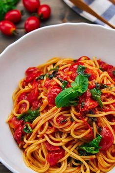 a white bowl filled with pasta and tomatoes on top of a table next to fresh basil