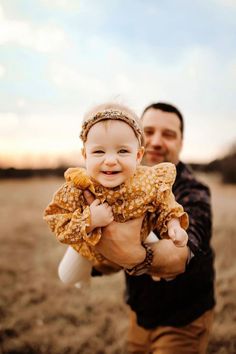 a man holding a baby in his arms while he smiles at the camera with an open field behind him