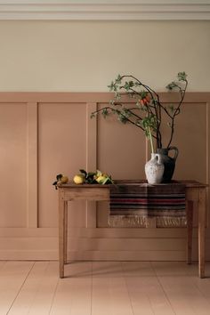 a wooden table topped with a white vase filled with flowers and fruit next to a wall