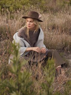 a woman sitting in the grass wearing a brown hat and blanket over her shoulders, looking off into the distance