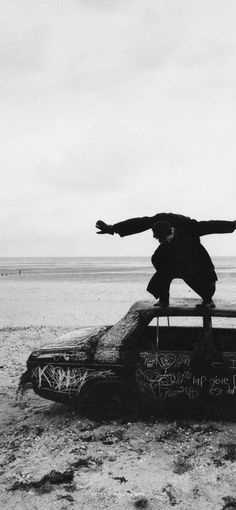 a man standing on top of an old car in the middle of nowhere with his arms outstretched