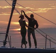 two people standing on the deck of a sailboat as the sun sets in the background