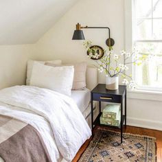 a white bed sitting under a window next to a wooden table with flowers on it