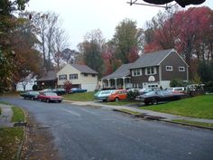 several cars parked on the side of a road in front of houses with fall foliage