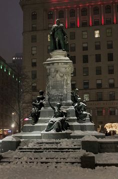 a statue in the middle of a snow covered park with buildings behind it at night