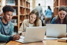 three students studying in the library on their laptops and taking notes while looking at books