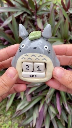 a person holding a ceramic animal clock in their hand, with plants behind the clock