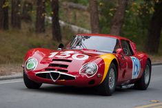 an old red sports car driving down the road in front of some tall trees and bushes