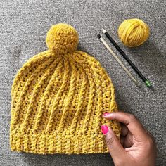 a hand holding a yellow knitted hat next to two crochet needles and a ball of yarn