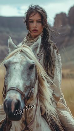 a woman riding on the back of a white horse in a desert area with mountains behind her