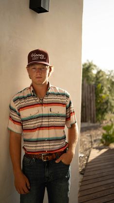 a man wearing a hat standing next to a wall with his hands in his pockets