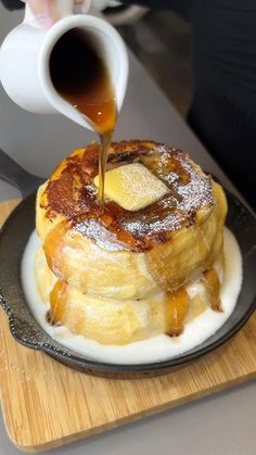 a person pouring syrup onto a stack of pancakes on a black plate with a wooden cutting board