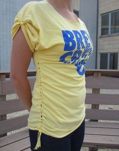 a woman wearing a yellow shirt with blue letters on the front and back, standing in front of a building