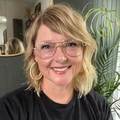 a woman wearing glasses and smiling for the camera in front of a window with potted plants