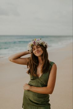 a pregnant woman wearing a flower crown on the beach with her hands behind her head