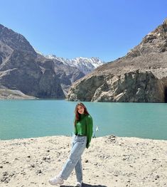 a woman standing on top of a sandy beach next to a mountain lake with snow capped mountains in the background