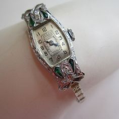 "Fabulous circa 1920s Bulova watch with four emeralds and two diamonds set into a lovely filigree curved tonneau-style case. The face has painted black number and blue steel hands. A decorative design on the face highlights the Bulova mark. The case also has filigree sides and the watch crown is engraved with a small flower.  The 14K white gold floral band has a fold over clasp and a safety chain. The watch case is marked Pat. Jun 10 1924 Bulova 14K. The band is also marked 14K .  The 15 jewel movement is marked Bulova Watch Co, and is marked 5AP, indicating the movement was made between 1926 and 1937.  This watch keeps great time as it's been recently serviced, and is in very good condition, with some light scratching .There is a deeper scratch in the crystal, which is seen under magnific Emerald Watch, Buyable Pins, Art Deco Watch, Antique Costume Jewelry, Bulova Watches, Vintage Timepiece, Great Falls, Safety Chain, Blue Steel