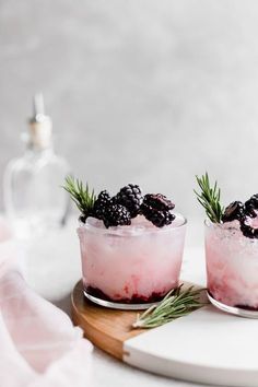 two glasses filled with ice and berries on top of a cutting board next to a bottle