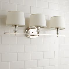 a bathroom light with three lamps on the wall and white tiled walls behind it,