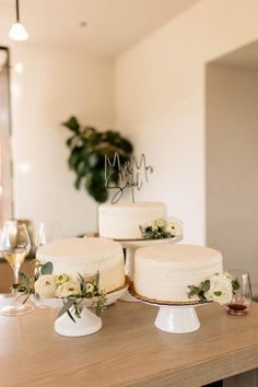 three white cakes sitting on top of a wooden table next to wine glasses and greenery