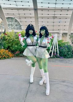 two women dressed in costumes standing next to each other on a sidewalk with flowers behind them