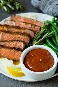 a plate with meat, mashed potatoes and green beans next to a small bowl of sauce