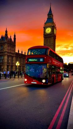 a red double decker bus driving down a street next to a tall tower with a clock on it's side