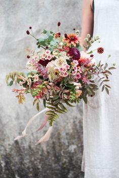 a woman holding a bouquet of flowers and greenery in front of a stone wall