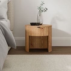 a small wooden table with a plant on it in front of a white wall and bed