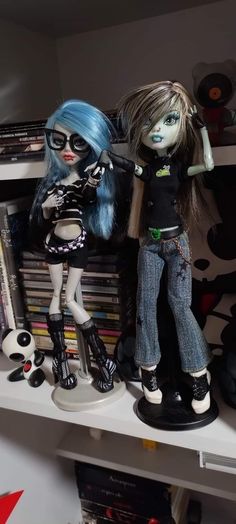 two dolls sitting on top of a white shelf next to books and cds in a room