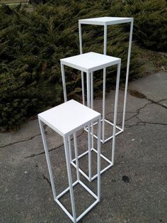 three white tables sitting next to each other on top of a cement ground with bushes in the background