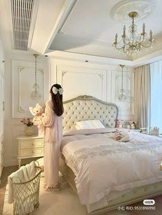 a woman standing in front of a bed with white furniture and chandelier hanging from the ceiling