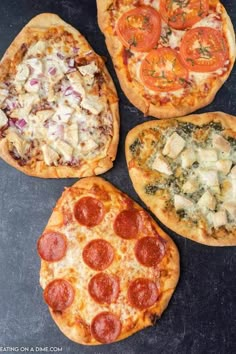 four pizzas with different toppings sitting on top of a black surface, each topped with cheese and tomatoes