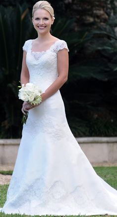 a woman in a wedding dress standing on the grass holding a bouquet and smiling at the camera