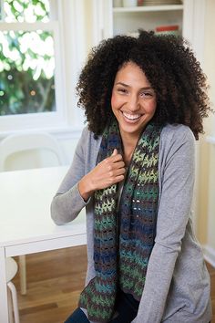 a woman sitting at a table smiling and wearing a knitted multicolored scarf