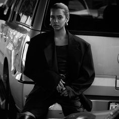 a woman sitting on the back of a car in front of a parked vehicle with her legs crossed