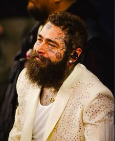 a man with tattoos on his face and beard sitting in front of another man wearing a white suit