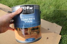 a hand holding a paint can in front of a wooden box with grass behind it