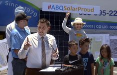 a man giving a speech at a podium surrounded by children in front of a solar panel