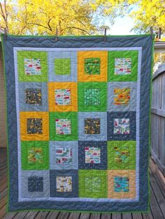 a green and yellow quilt hanging on a wooden deck next to a fence with trees in the background