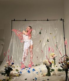 a woman standing in front of a white backdrop with flowers and petals on the ground